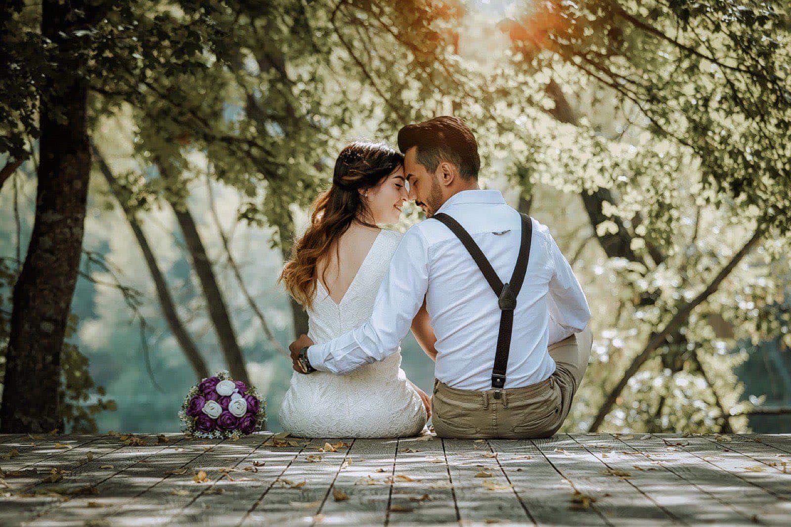 Pareja de novios sentada de espaldas en un muelle de madera bajo los árboles, frente con frente, con el ramo a un lado
