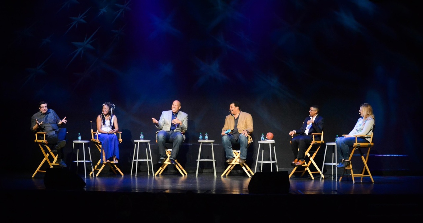 Seis personas conversando sentadas en banquillos sobre un escenario oscuro, iluminadas con luz azul de teatro