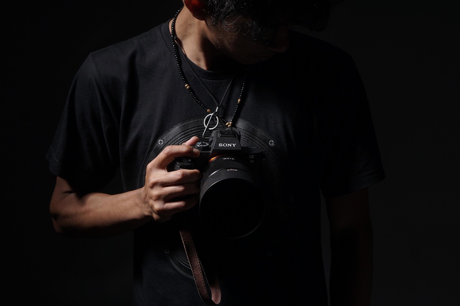 Fotógrafo con camiseta negra sosteniendo una cámara sin espejo colgada al cuello, sobre fondo oscuro de estudio