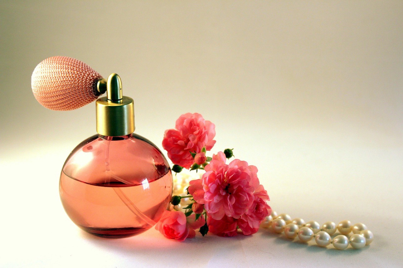 Frasco de perfume de vidrio rosa con perilla de atomizador, acompañado de rosas y un collar de perlas sobre fondo neutro