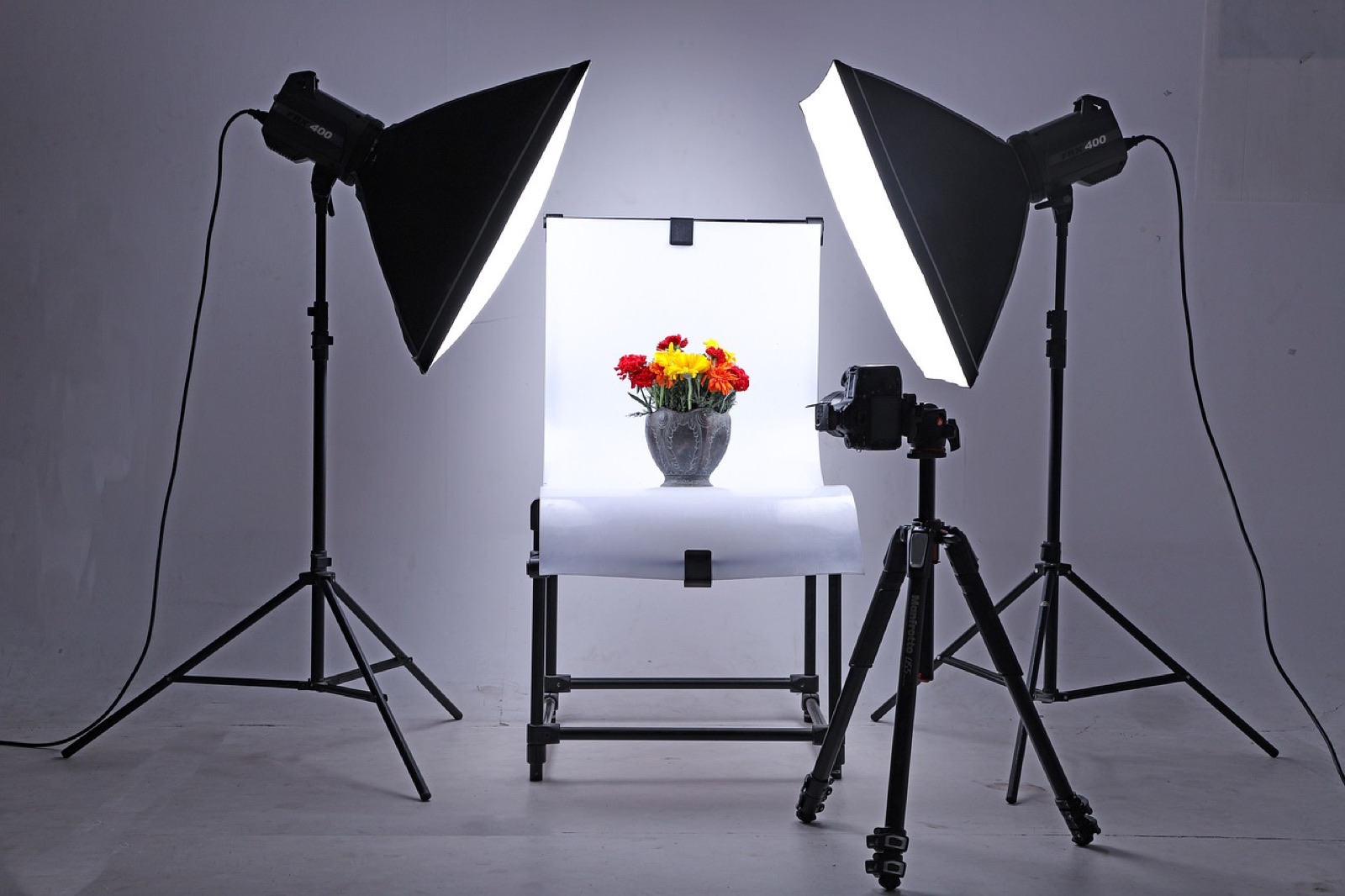Mesa de fotografía de producto con fondo blanco y un jarrón de flores, iluminado por dos softboxes, con una cámara en tripié apuntando al montaje