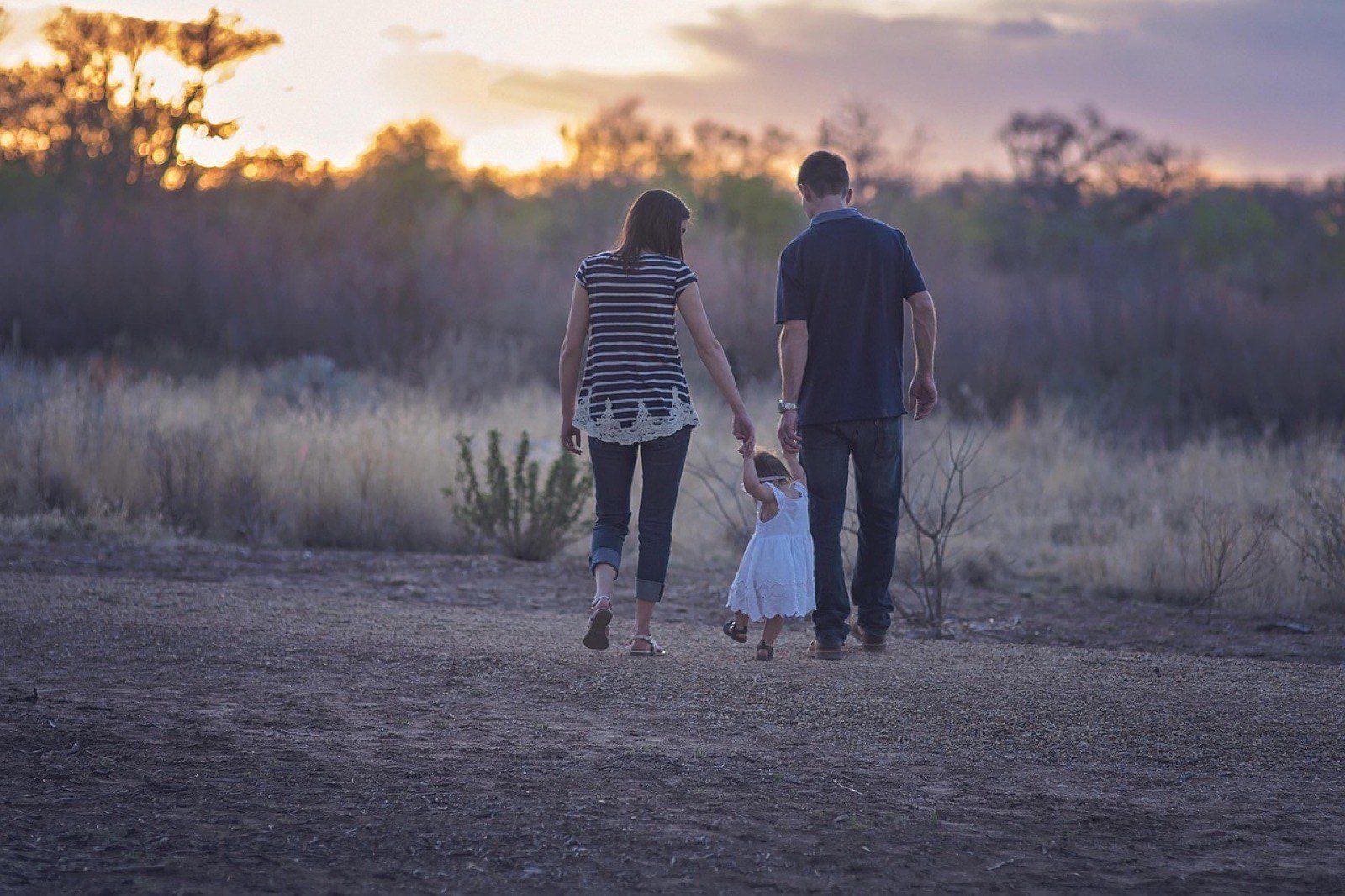 Padre y madre caminando de espaldas por un campo al atardecer, llevando de la mano a una niña pequeña con vestido blanco